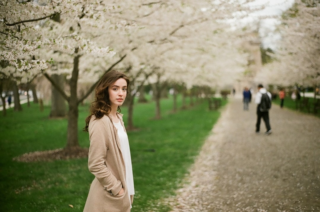 On a college campus in springtime, a woman with dark brown hair is looking at the camera. Trees with white blossoms are in the background.
